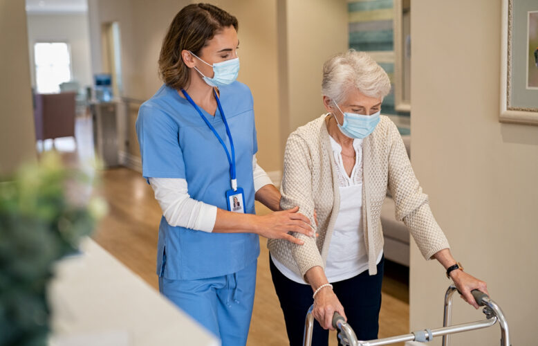 A healthcare worker wearing blue scrubs and a surgical mask helps an elderly woman using a walker.