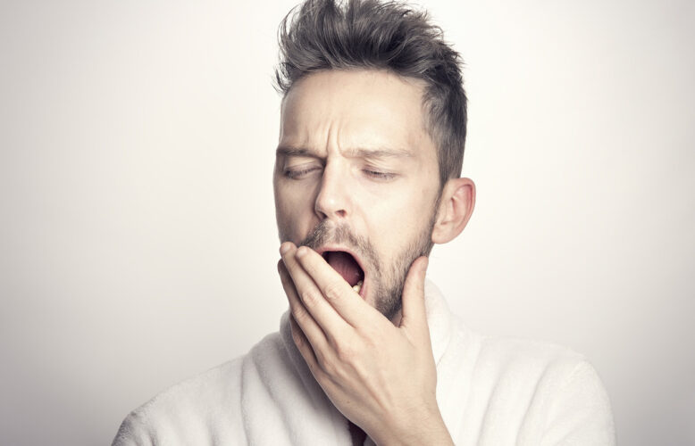 Young man with dark hair is yawning. His left hand is covering his open mouth.