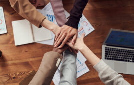 Hands in, stacked atop each other in solidarity, with a business office table in the background.