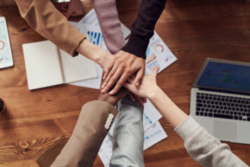 Hands in, stacked atop each other in solidarity, with a business office table in the background.
