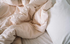 Photo of a rumpled, unmade bed with blankets, sheets and pillows. The blanket is a light cream color.